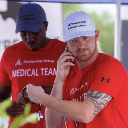 Two members of a medical team are pictured under a shade at one of the aid stations. They are both wearing red shirts with "Ascension Seton MEDICAL TEAM" written on them. One is holding a stethoscope and talking on a phone; the other is looking down and adjusting the stethoscope. Austin Marathon Half Marathon & 5K